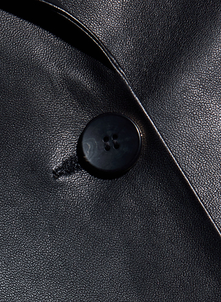 Close-up of a black button sewn onto smooth, shiny black leather fabric from the Chamberlain Leather Blazer—showcasing the classic style with a visible buttonhole and neatly folded edge of the material.
