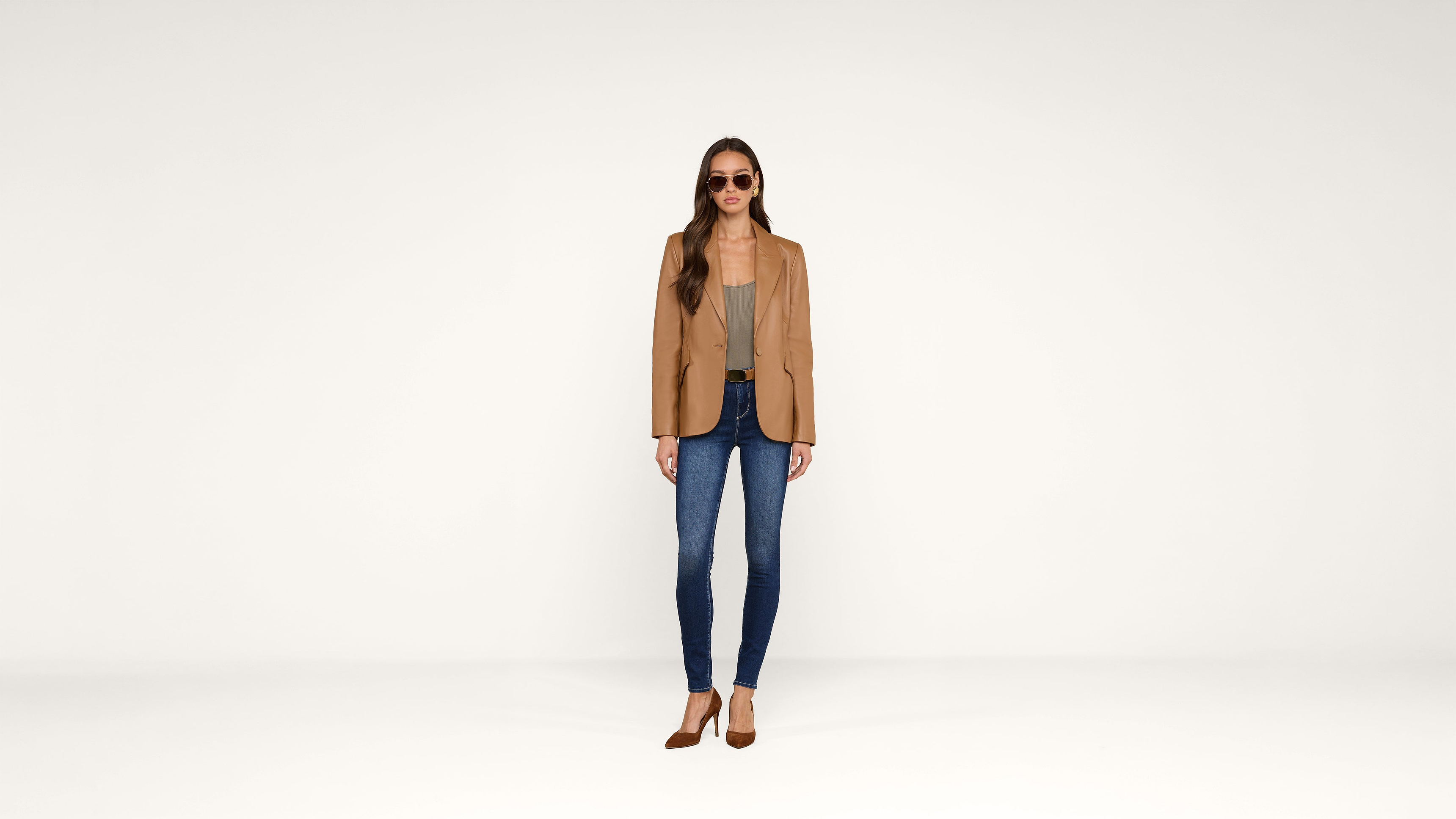 A woman wearing sunglasses, a tan blazer, gray top, blue jeans, and brown heels stands against a plain white background.