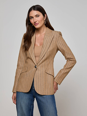 A woman with long brown hair wears the L’AGENCE Chamberlain Blazer—a tan, single-breasted pinstripe style—over a beige top and blue jeans, standing against a plain light gray background.
