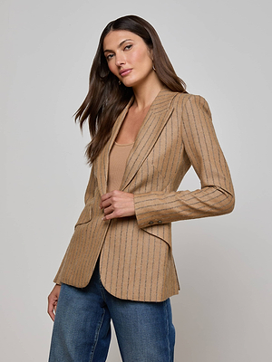A woman with long brown hair wears the Chamberlain Blazer by L'AGENCE over a beige top and blue jeans, posing against a plain light gray background.