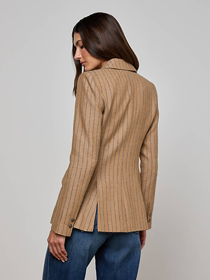 A woman with long brown hair models the L'AGENCE Chamberlain Blazer, a tan cotton-blend pinstripe jacket, paired with blue jeans. She stands with her back to the camera, looking over her left shoulder against a plain background.