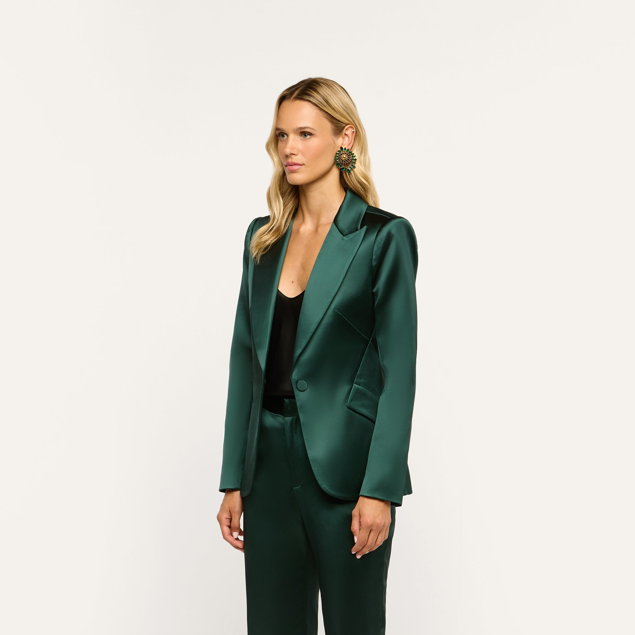 A person wearing a forest green Chamberlain Satin Blazer and matching trousers over a black top, accented with a large, round green and gold earring.