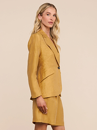 A woman with blonde hair stands sideways, looking at the camera, against a plain light background, wearing the Chamberlain Linen-Blend Blazer in mustard yellow with a matching skirt.