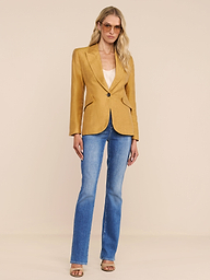 A woman stands against a plain white background, looking straight ahead with a neutral expression. She is wearing the Chamberlain Linen-Blend Blazer in gold, styled over a light top, blue jeans, and brown sandals.