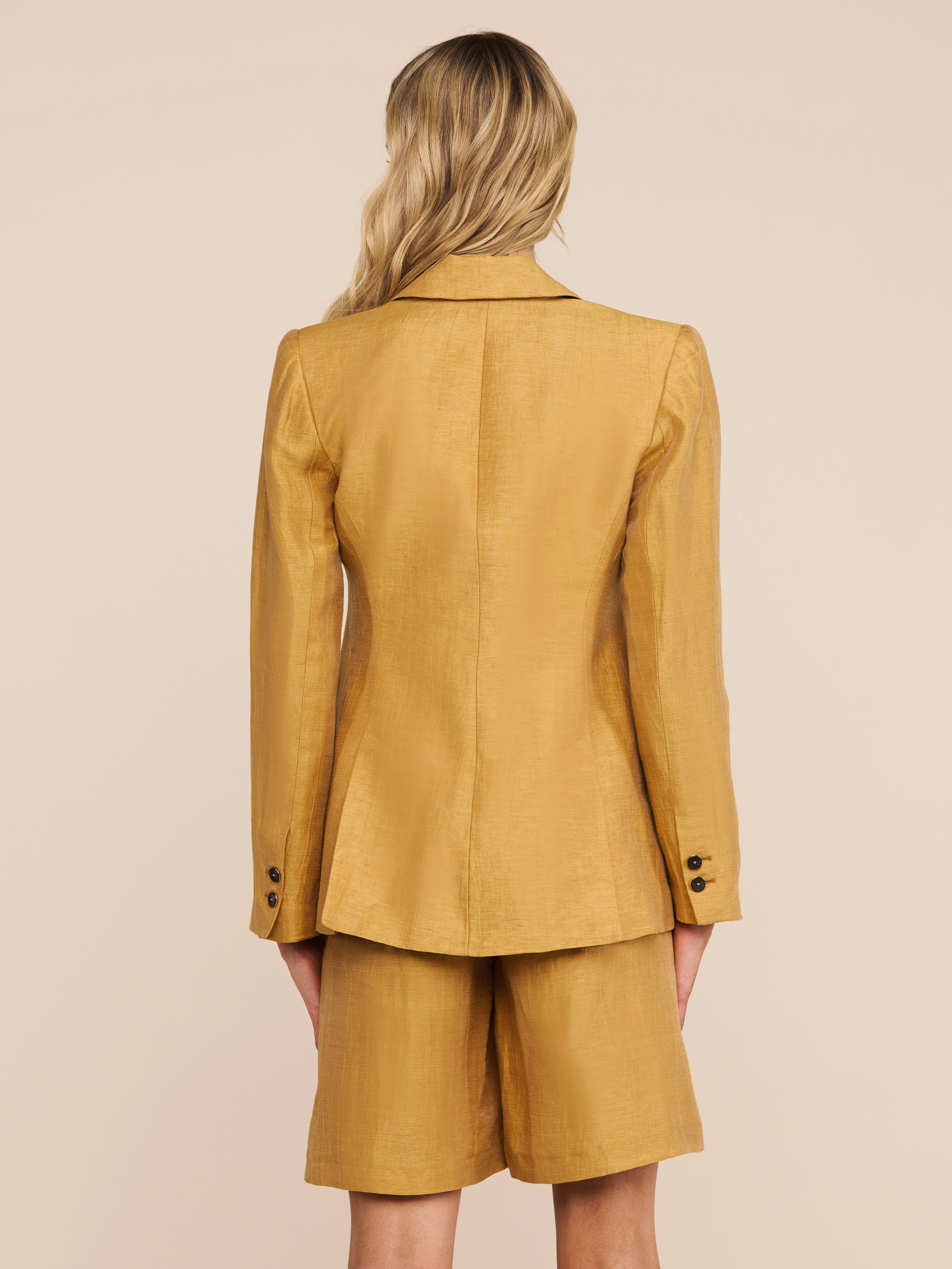 A person with wavy blonde hair is standing with their back to the camera, wearing the Chamberlain Linen-Blend Blazer in gold and matching knee-length shorts. The background is plain and light-colored.