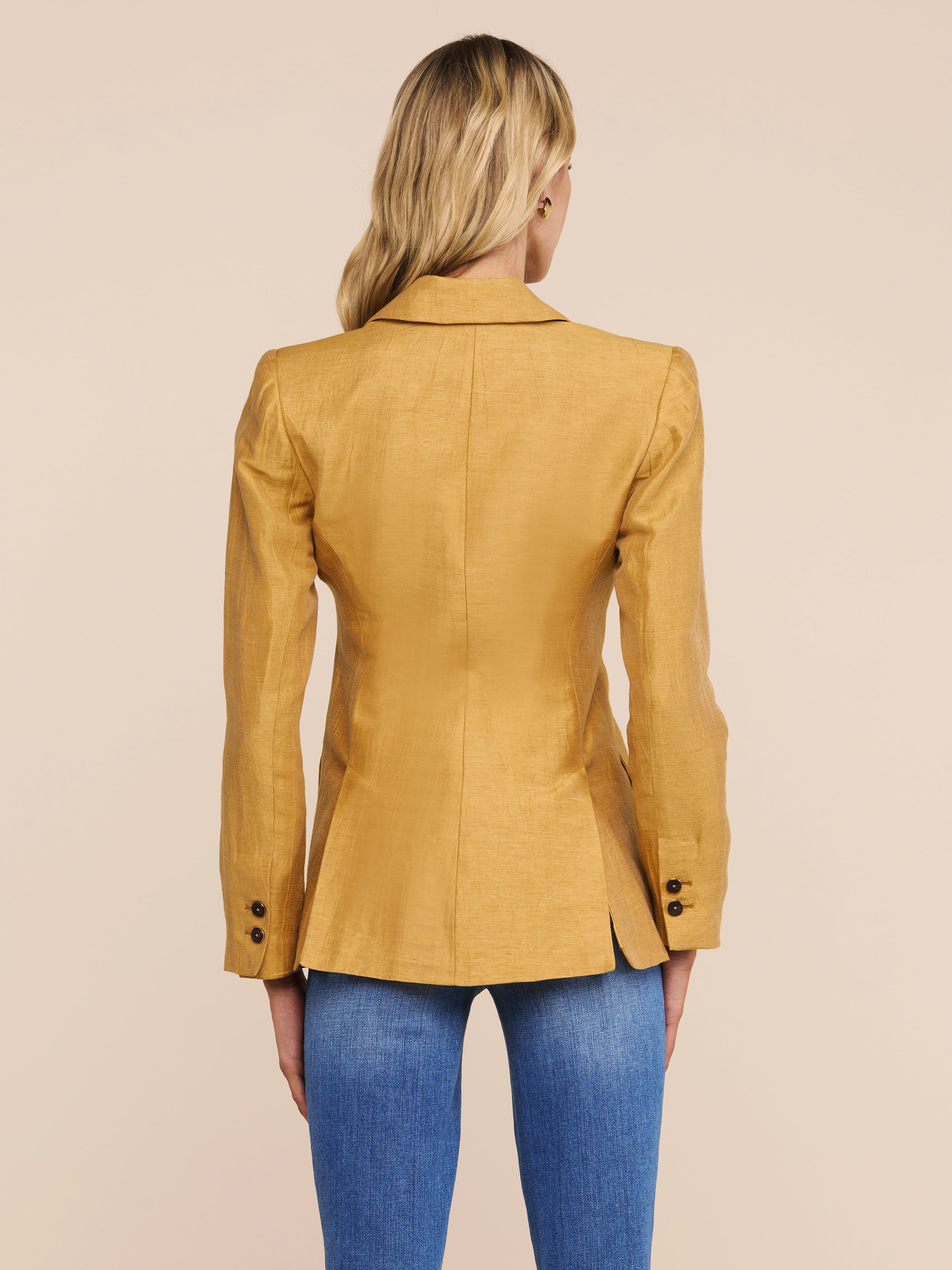 A woman with long blonde hair, seen from behind, wears the Chamberlain Linen-Blend Blazer in mustard yellow with buttoned sleeves and blue jeans, standing against a plain light background.