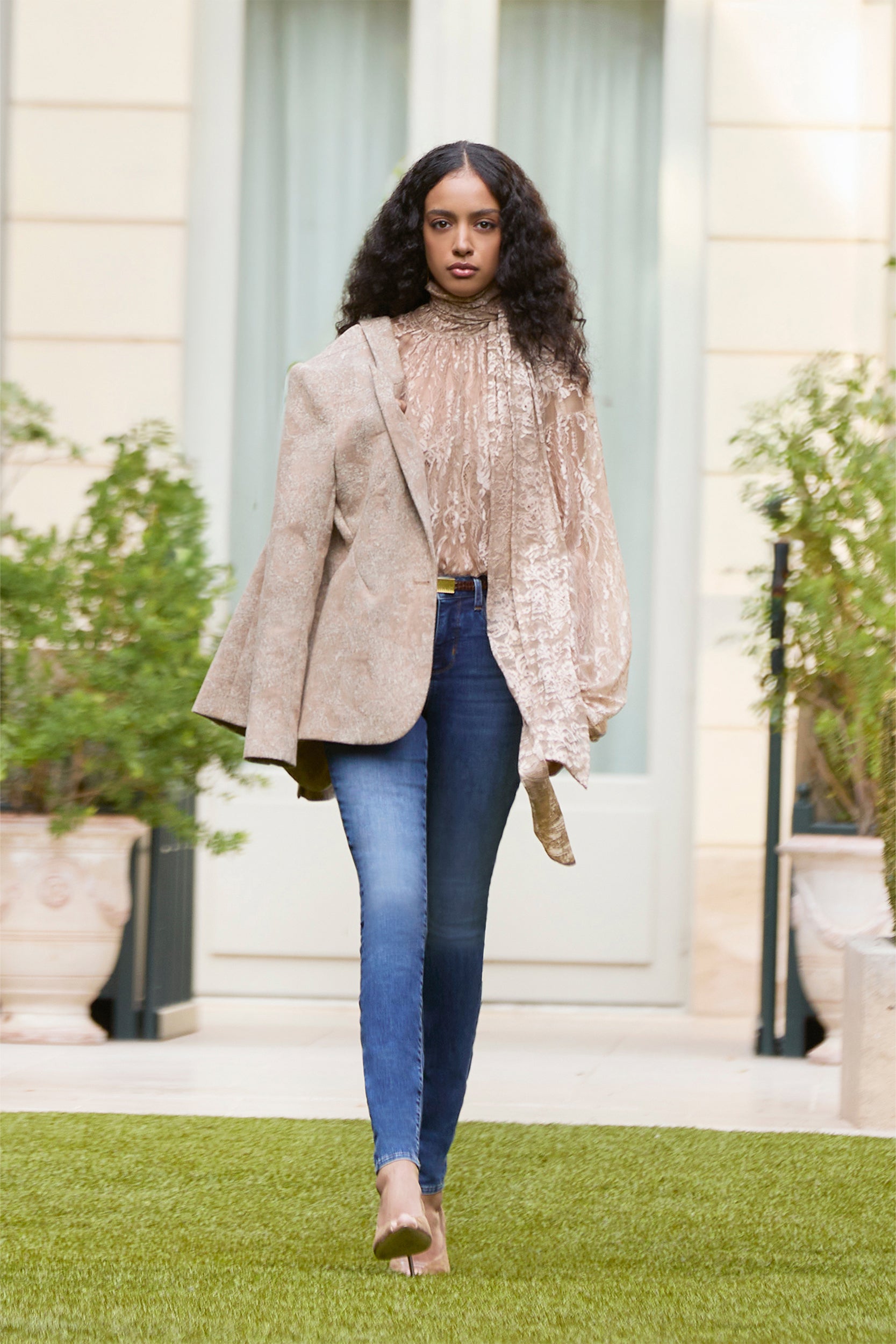 A woman with curly hair models a beige asymmetrical blazer over a shiny blouse and blue jeans, strolling outdoors on grass amid potted plants and a modern building in the background.