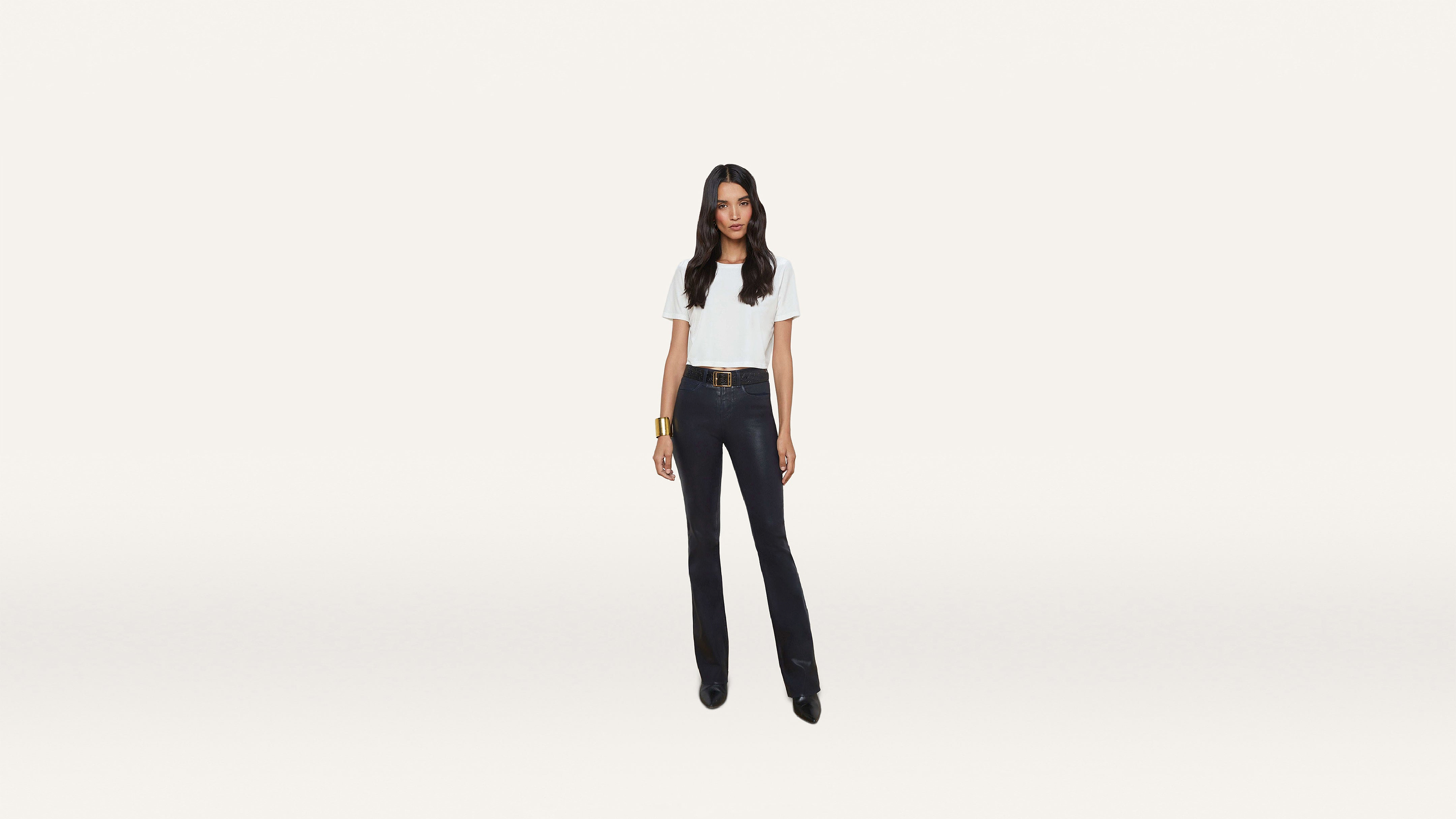 A woman stands against a plain white background, wearing a crisp white t-shirt, dark pants, black shoes, and a stylish bracelet.