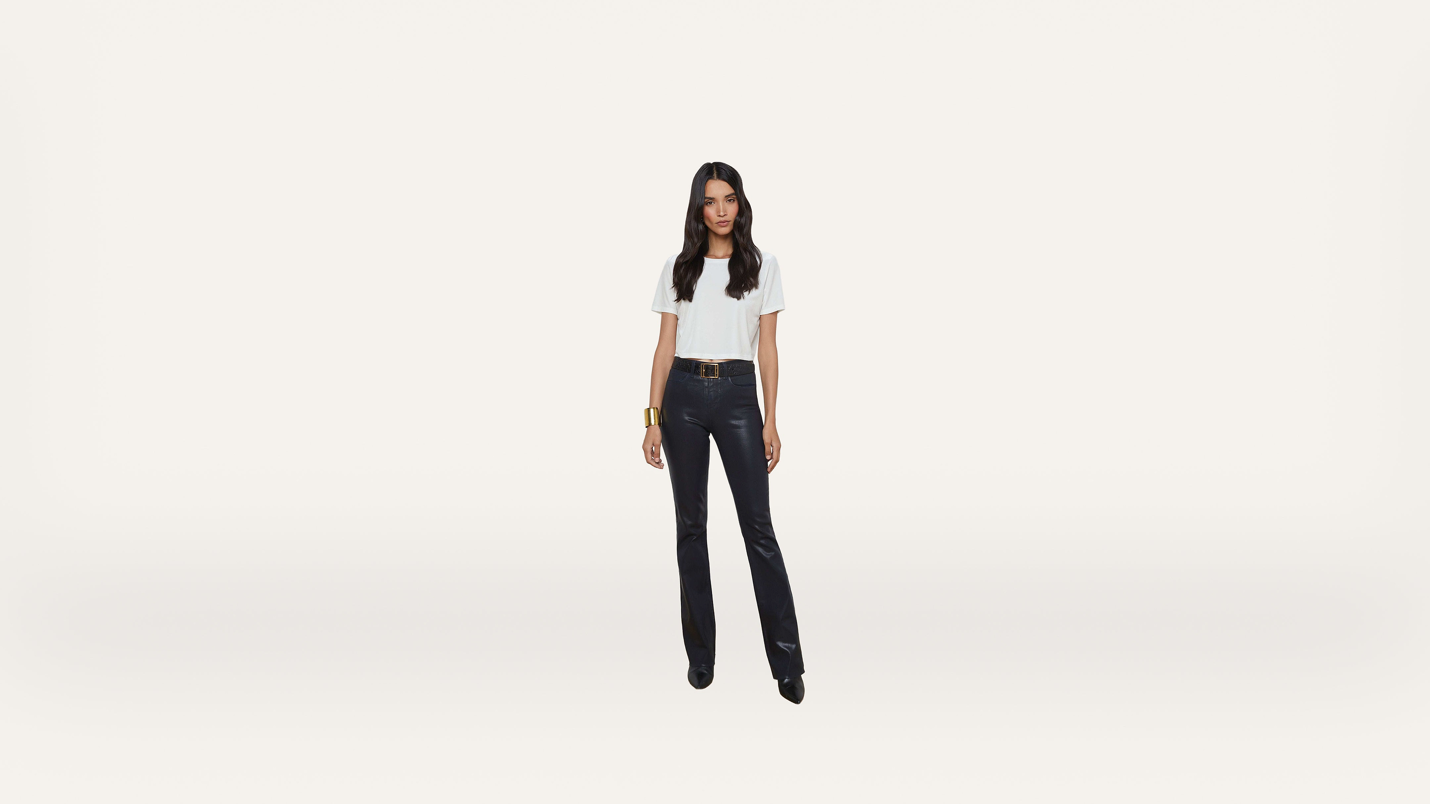 A woman stands facing forward, wearing a white short-sleeve top, black pants, and black shoes. She has long, dark hair and a gold bracelet, posing against a plain white background.
