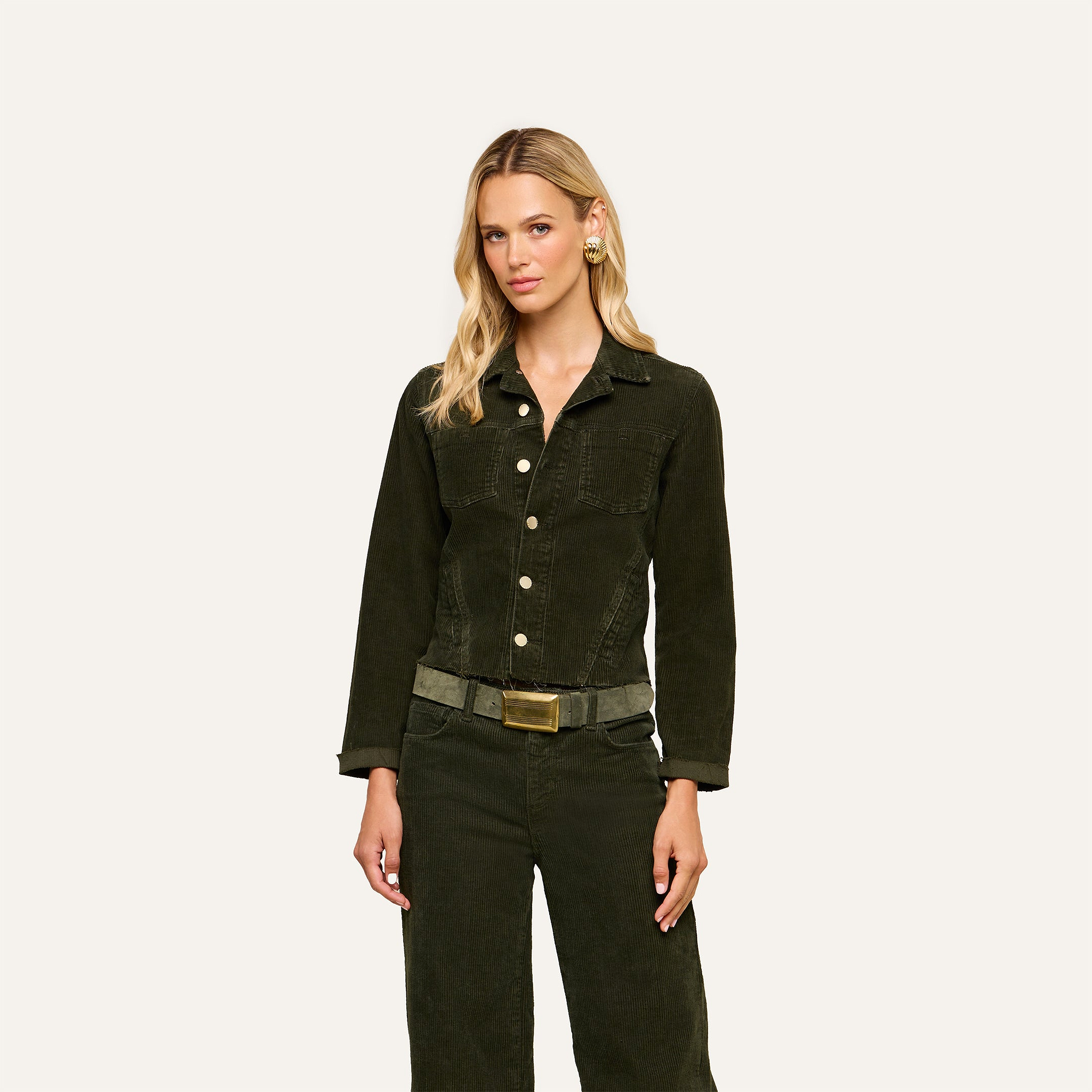 A woman with long blonde hair wears the Janelle Corduroy Jacket and matching green pants with a belt, standing against a plain light background.