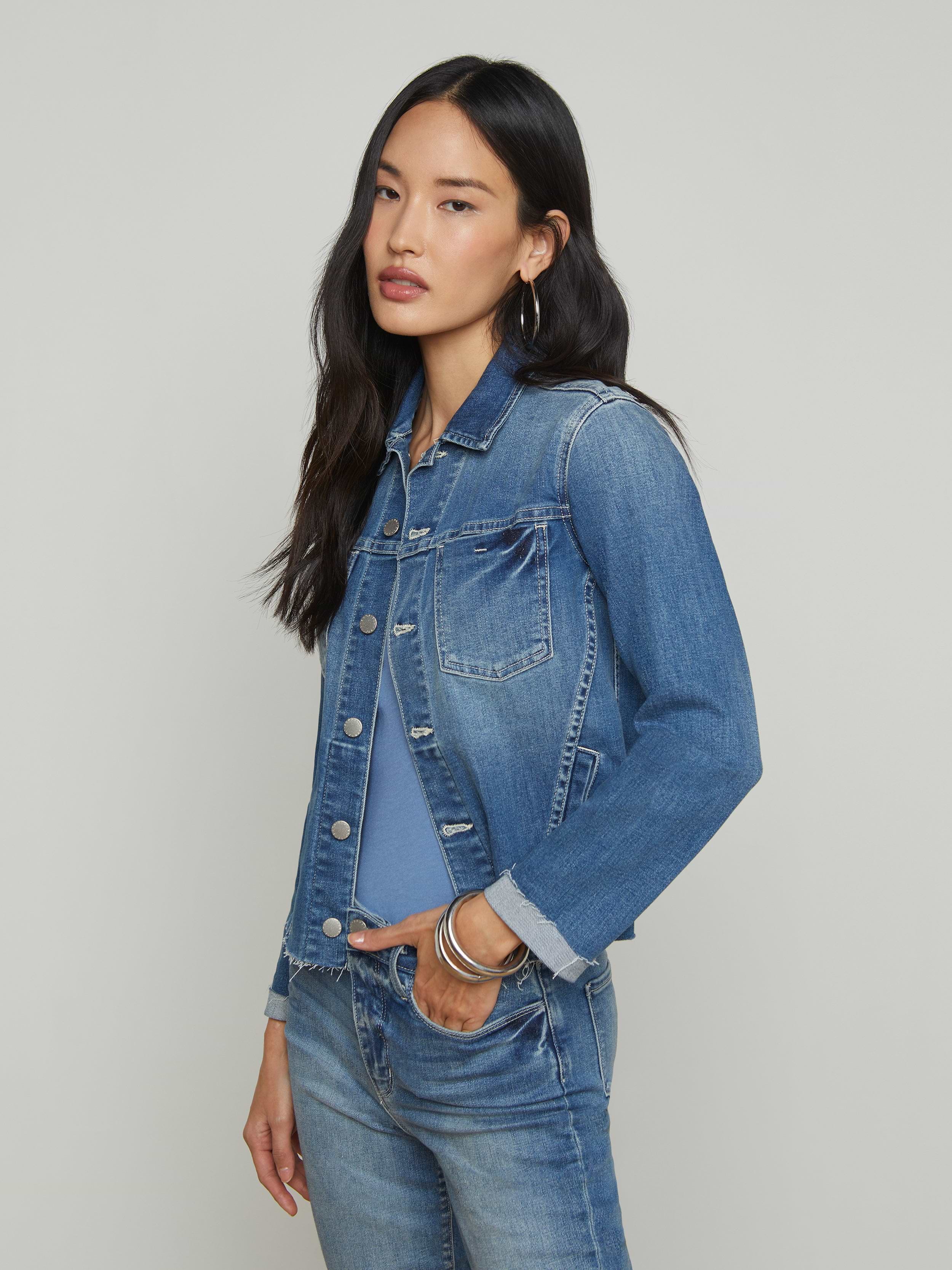 A woman with long dark hair wearing a blue denim jacket over a blue shirt and blue jeans stands with one hand in her pocket against a plain light background.