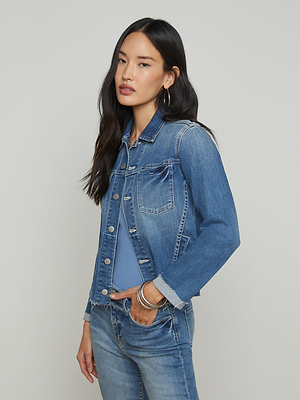 A woman with long dark hair wearing a blue denim jacket over a blue shirt and blue jeans stands with one hand in her pocket against a plain light background.