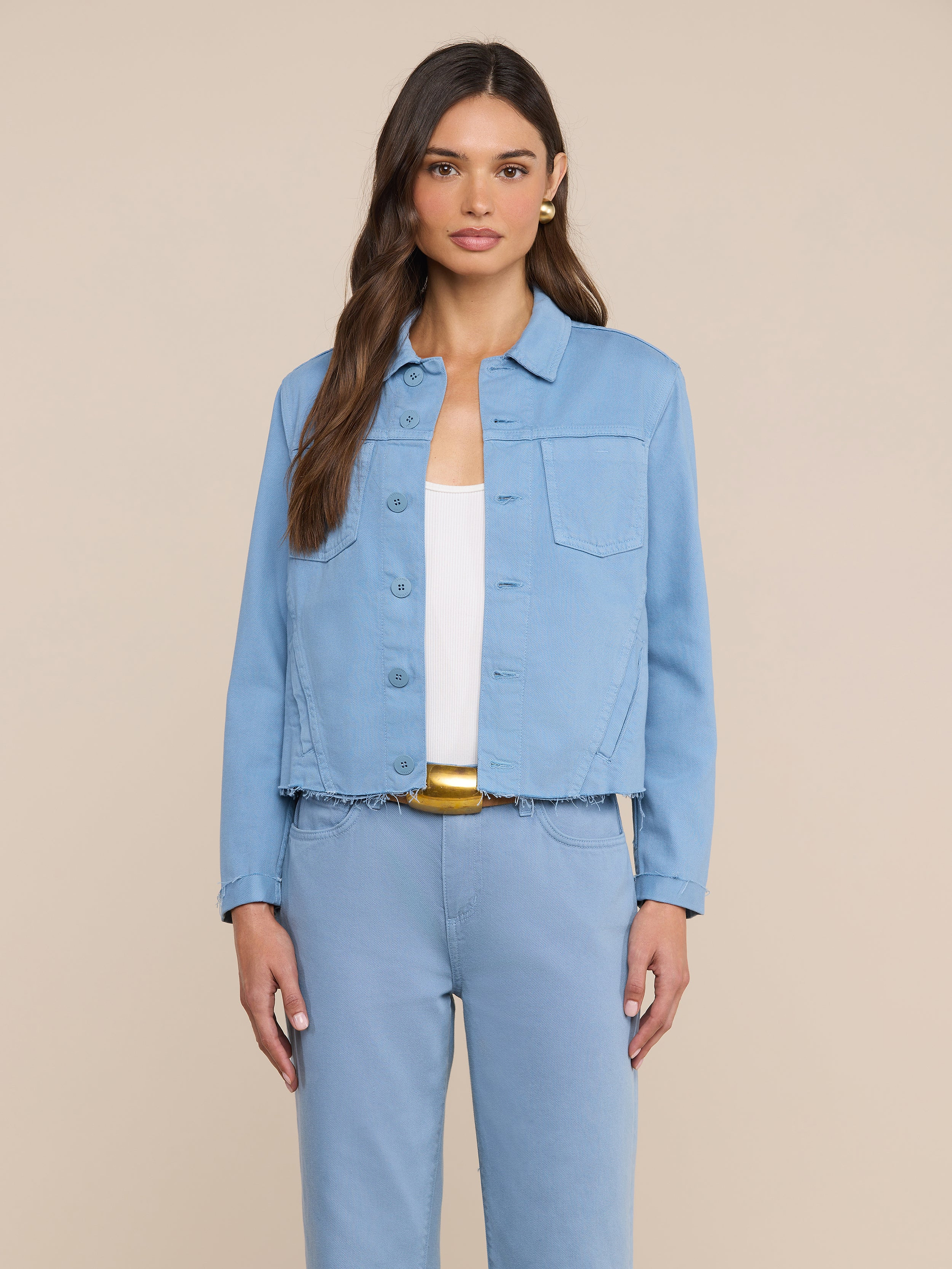 Una donna dai lunghi capelli castani indossa una giacca Janelle in denim blu dal taglio aderente e pantaloni abbinati sopra un top bianco, in piedi davanti a uno sfondo beige a tinta unita.