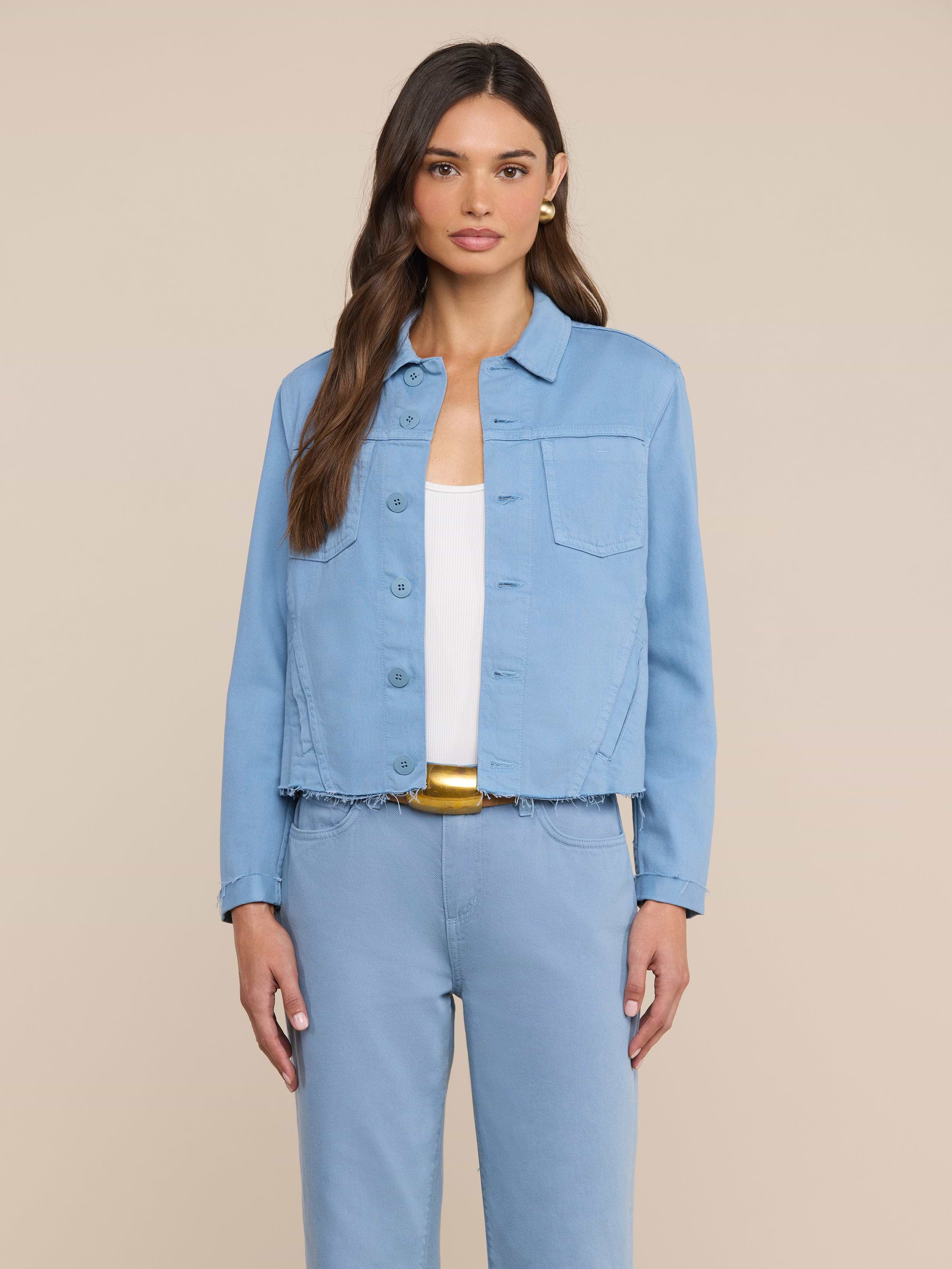 Una donna dai lunghi capelli castani indossa una giacca Janelle in denim blu dal taglio aderente e pantaloni abbinati sopra un top bianco, in piedi davanti a uno sfondo beige a tinta unita.