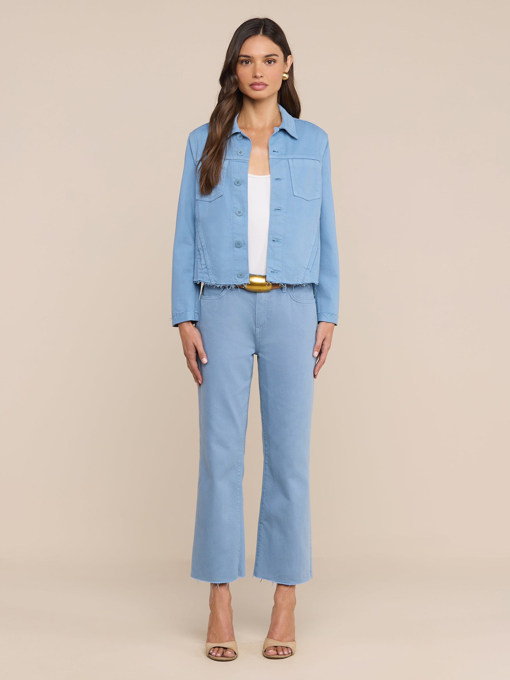 Una donna è in piedi, rivolta verso la fotocamera, su uno sfondo beige; indossa i jeans corti a gamba larga Wanda, abbinati a una giacca di jeans azzurra, un top bianco e scarpe con tacco aperte in punta di colore beige.