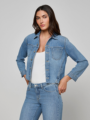 A woman with long dark hair models the Janelle Jean Jacket—a pinstriped denim style—over a white top and matching striped jeans, standing against a plain light gray background.