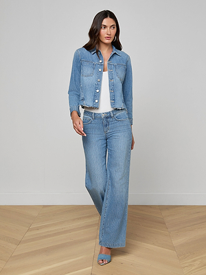 A woman with long dark hair wears the Janelle Jean Jacket and matching wide-leg jeans over a white top, paired with blue open-toe heels, as she walks on a light wood floor against a plain light gray wall.