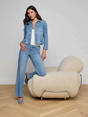 A woman models the Janelle Jean Jacket and matching jeans, paired with a white top and blue heels. She stands confidently with one foot on a cream chair in a minimalist room with wooden floors, her long dark hair completing the look.