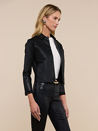 A woman with long blonde hair models the Janelle Coated Jean Jacket over a white top and black leather pants, standing in a relaxed pose against a plain beige background and wearing large round earrings.