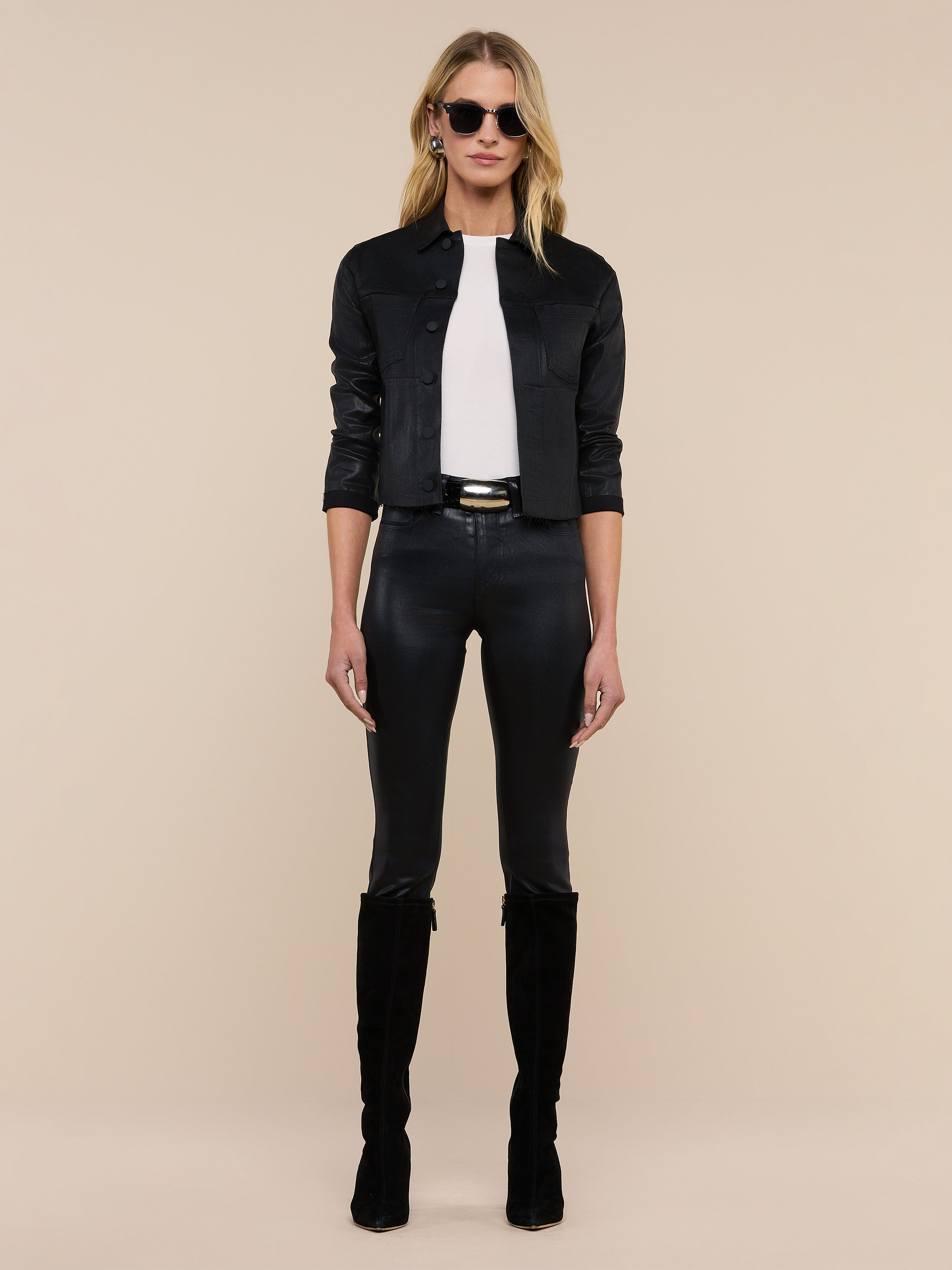 A woman stands facing forward in sunglasses, the Janelle Coated Jean Jacket, a white top, black fitted pants, and knee-high black boots against a plain beige background.