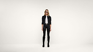 A woman stands against a plain white background, wearing sunglasses, a black jacket, a white shirt, black pants, and knee-high black boots.