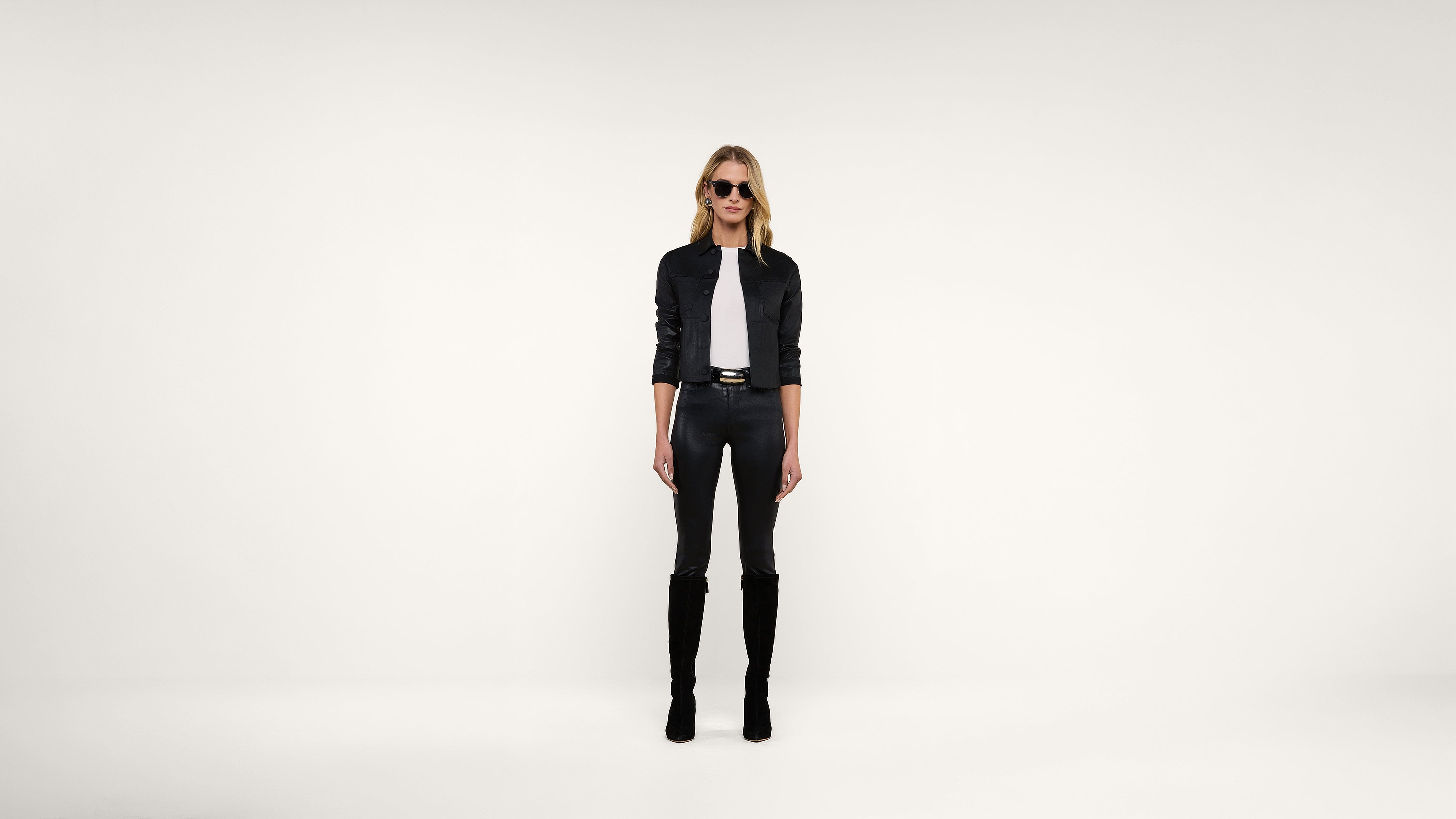 A woman stands against a plain white background, wearing sunglasses, a black jacket, a white shirt, black pants, and knee-high black boots.