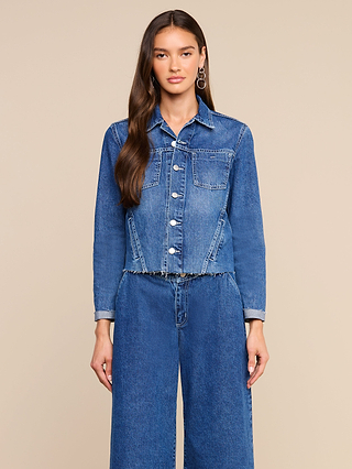 A woman with long brown hair stands against a beige background, wearing the Janelle Jean Jacket—a medium wash denim jacket with a raw hem—paired with wide-leg blue jeans. She looks directly at the camera.