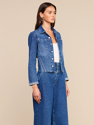 A woman with long brunette hair and large hoop earrings wears the Janelle Jean Jacket in a medium wash over a white top, paired with wide-leg denim pants. She stands against a neutral beige background.