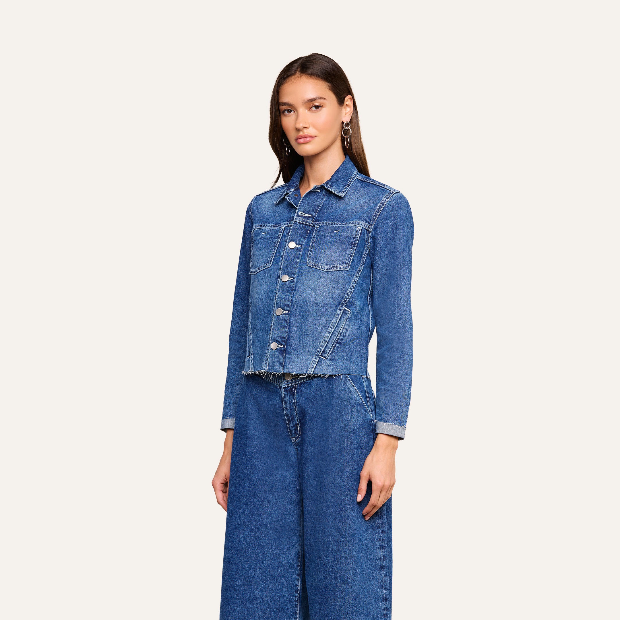 A woman stands facing forward, wearing the Janelle Jean Jacket in a medium wash with front pockets and a slim fit, paired with matching wide-leg blue jeans against a plain light background.