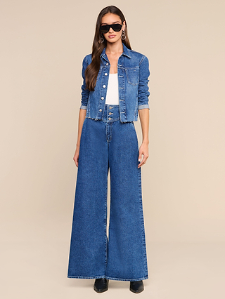 Woman wearing sunglasses, hoop earrings, the Janelle Jean Jacket in a medium wash, a white top, and high-waisted wide-leg denim jeans, standing against a plain beige background.