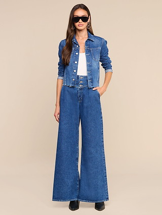 A woman wearing sunglasses, a white shirt, the Janelle Jean Jacket in a slim fit, and high-waisted wide-leg medium wash denim jeans stands with one hand in her pocket against a beige background.