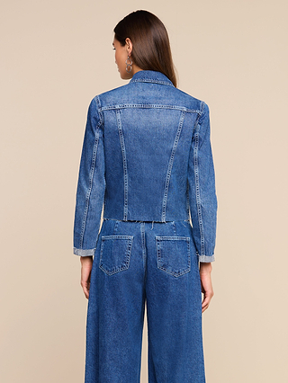 A woman with straight brown hair models the Janelle Jean Jacket, a slim fit denim jacket in a medium wash, along with wide-leg jeans, shown from the back against a plain beige background.