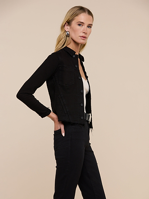 A woman with long blonde hair models the Janelle Jean Jacket over a white top and black pants, standing against a plain beige background with one hand on her hip and gazing slightly to the side.