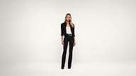 A woman stands in a minimal, white studio wearing a black jacket, white top, black pants, and black shoes, posing with a relaxed posture and a neutral expression.