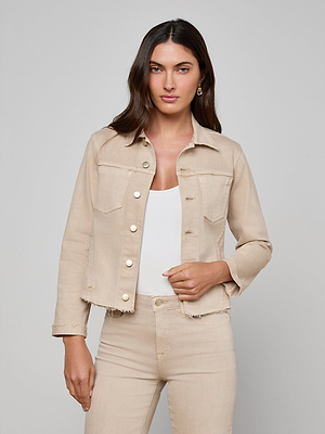 A woman with long brown hair wears the Janelle Jean Jacket in beige, styled over a white top and matching beige jeans. She stands against a plain light gray background, looking at the camera with a neutral expression.