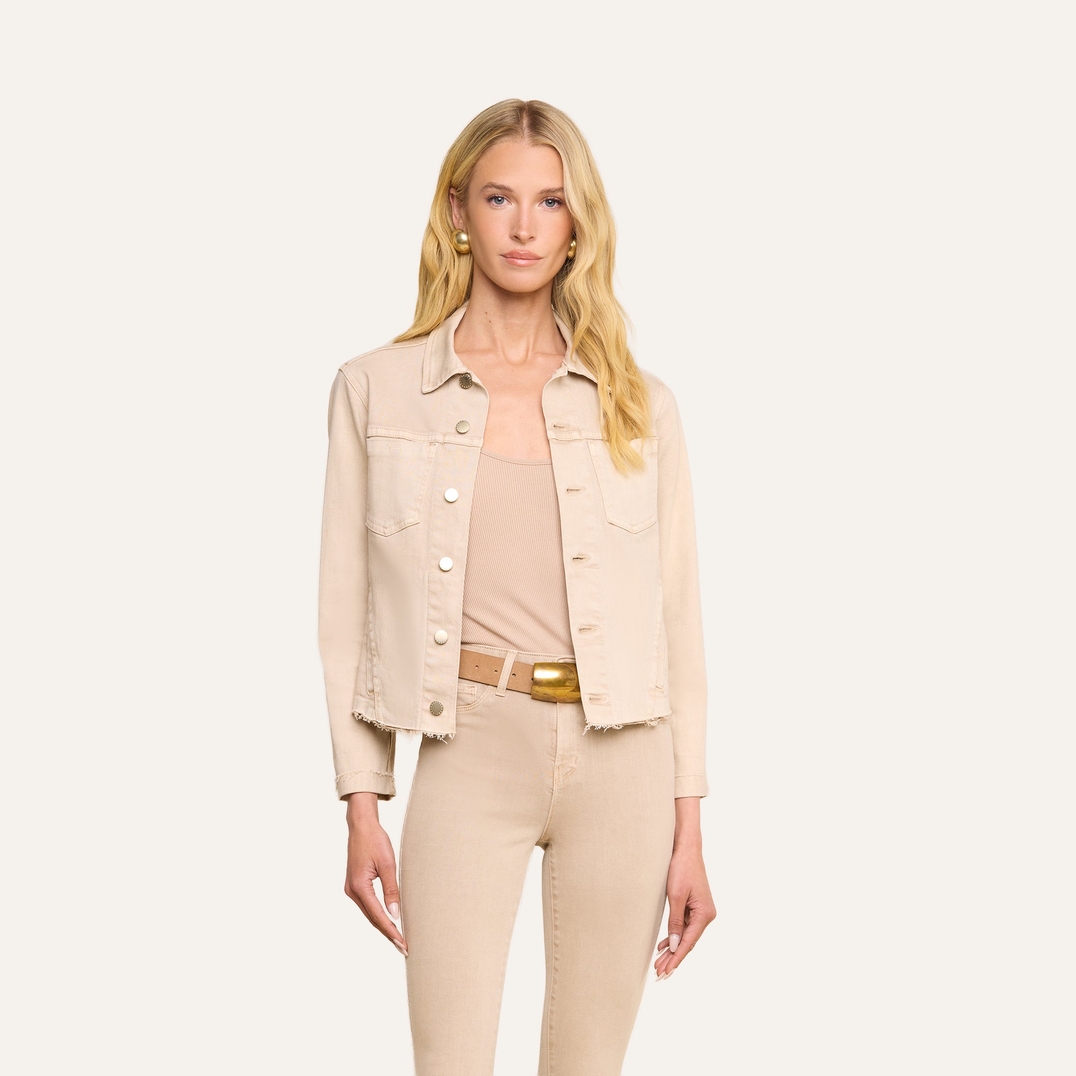 Une femme aux longs cheveux blonds porte la veste « Janelle Jean » de couleur beige avoine, associée à un débardeur assorti, un pantalon taille haute, une ceinture marron et de grandes boucles doreilles en perles, le tout sur un fond beige.