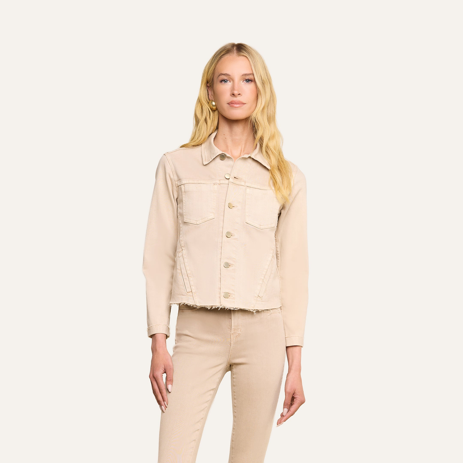 Una donna dai lunghi capelli biondi indossa la giacca Janelle Jean e i pantaloni abbinati in beige avena, dal taglio femminile. È in piedi, rivolta in avanti, su uno sfondo beige a tinta unita, con una mano lungo il fianco e unespressione neutra.