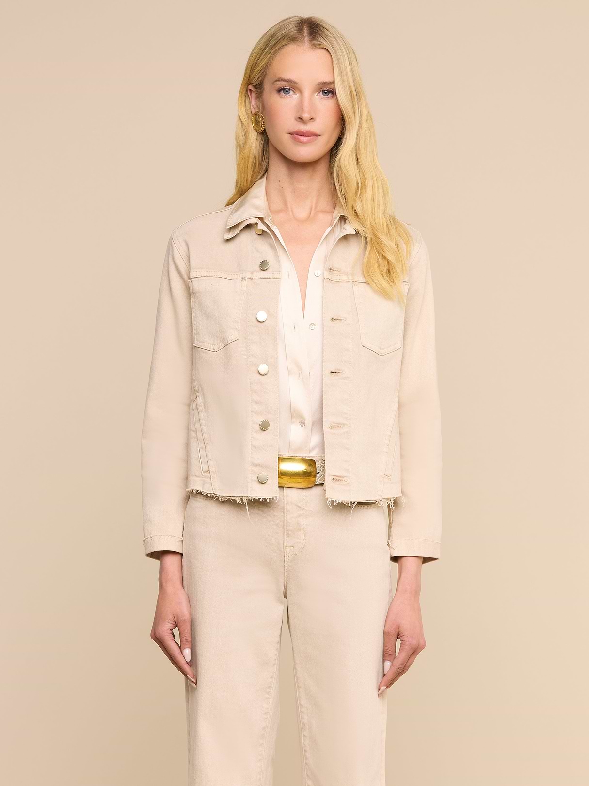 Una donna dai lunghi capelli biondi guarda in avanti, indossa la giacca Janelle Jean color beige avena sopra una camicia bianca e pantaloni beige abbinati, su uno sfondo beige a tinta unita.