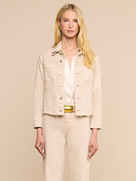 Une femme aux longs cheveux blonds, tournée vers lavant, porte la veste « Janelle Jean » de couleur beige avoine par-dessus une chemise blanche et un pantalon beige assorti, sur fond beige uni.