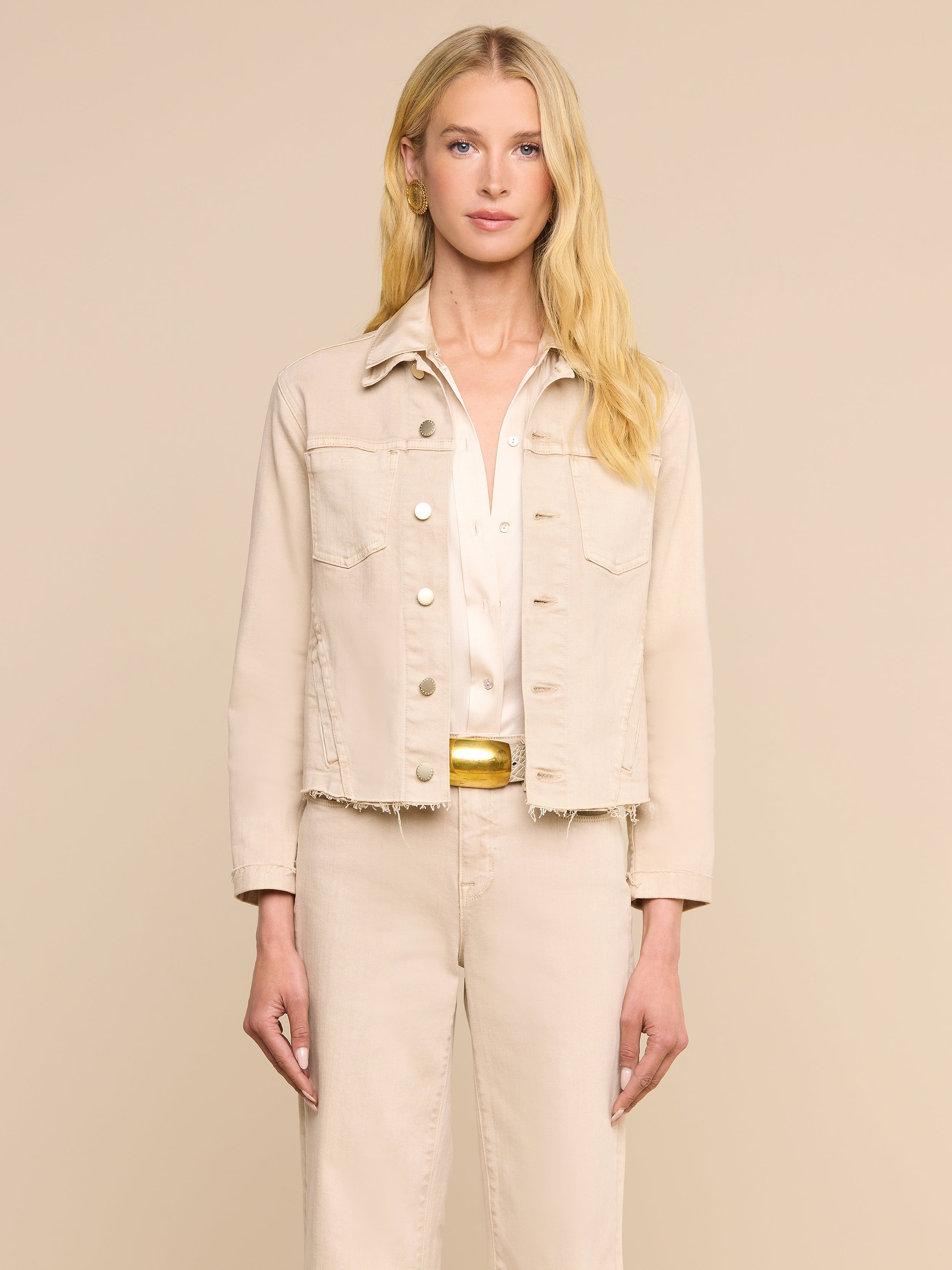 Une femme aux longs cheveux blonds, tournée vers lavant, porte la veste « Janelle Jean » de couleur beige avoine par-dessus une chemise blanche et un pantalon beige assorti, sur fond beige uni.