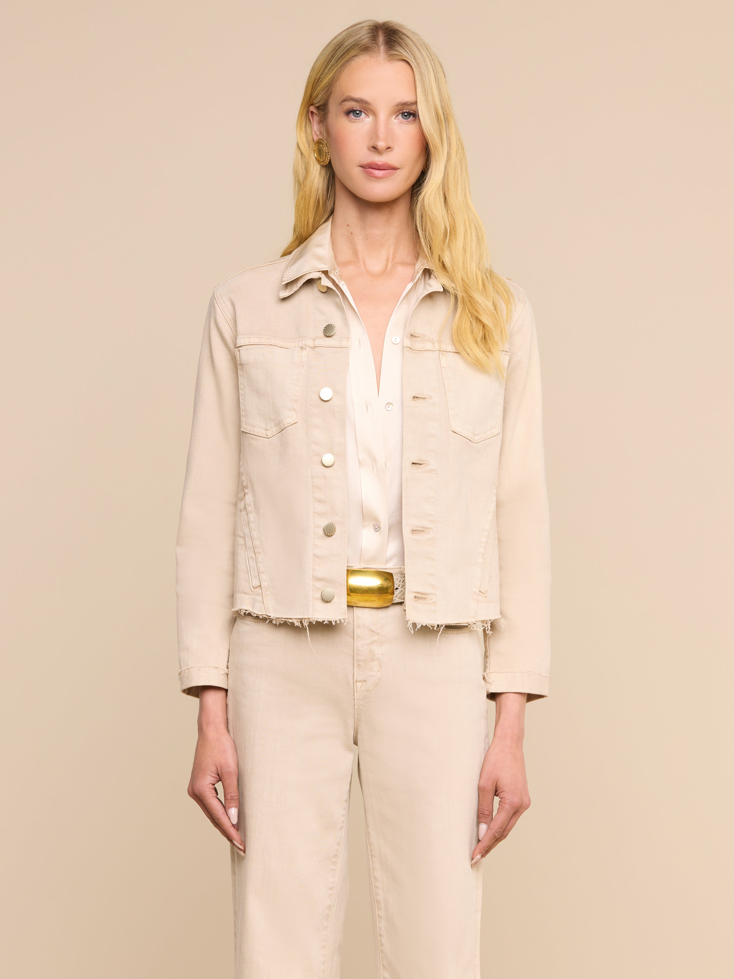 Une femme aux longs cheveux blonds, tournée vers lavant, porte la veste « Janelle Jean » de couleur beige avoine par-dessus une chemise blanche et un pantalon beige assorti, sur fond beige uni.