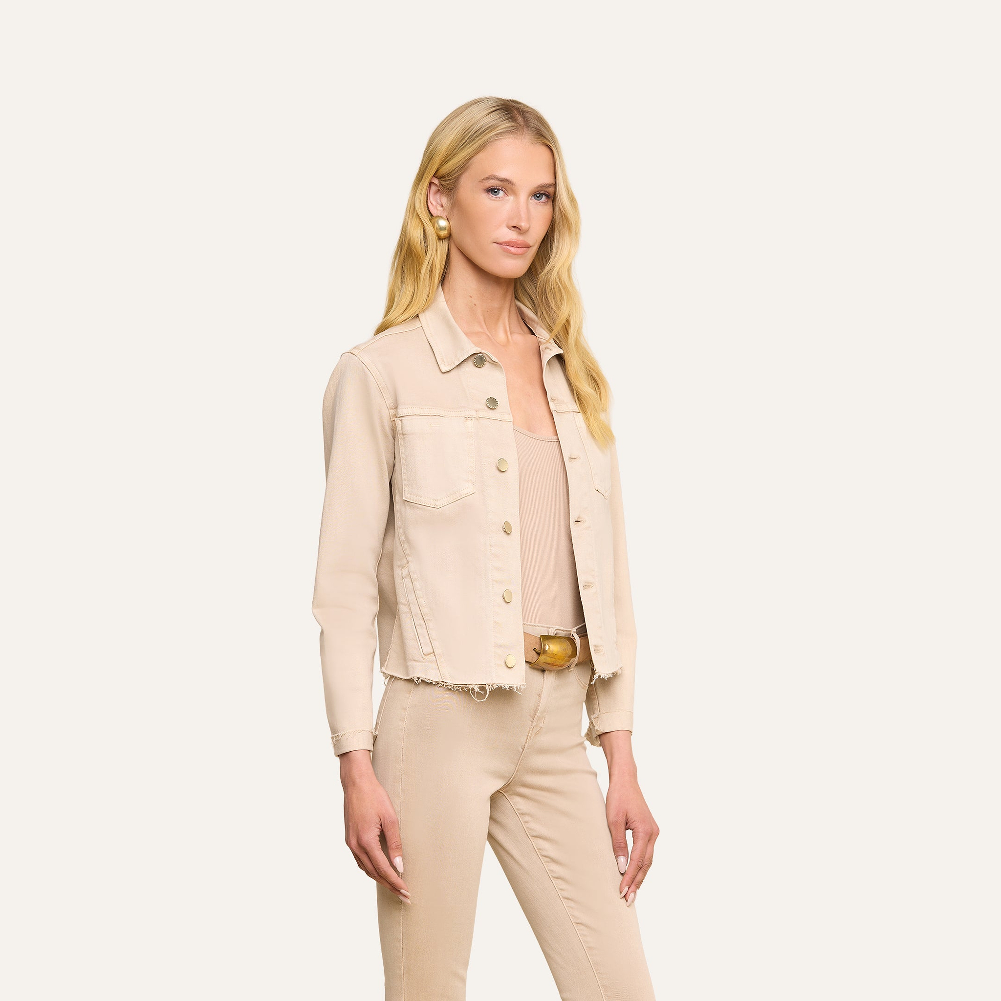 Une femme aux longs cheveux blonds se tient de profil, vêtue de la veste « Janelle Jean » — une veste en jean de couleur beige avoine à la coupe féminine —, assortie dun pantalon en jean stretch de la même couleur, dun haut beige clair et de boucles doreilles créoles dorées, sur un fond beige.