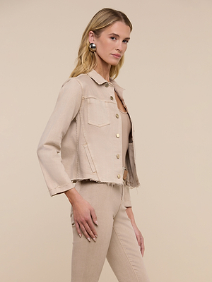 A woman with long blonde hair wears the Janelle Jean Jacket in oatmeal beige with matching feminine-fit pants, standing sideways against a neutral background. She has large circular earrings and a calm expression.