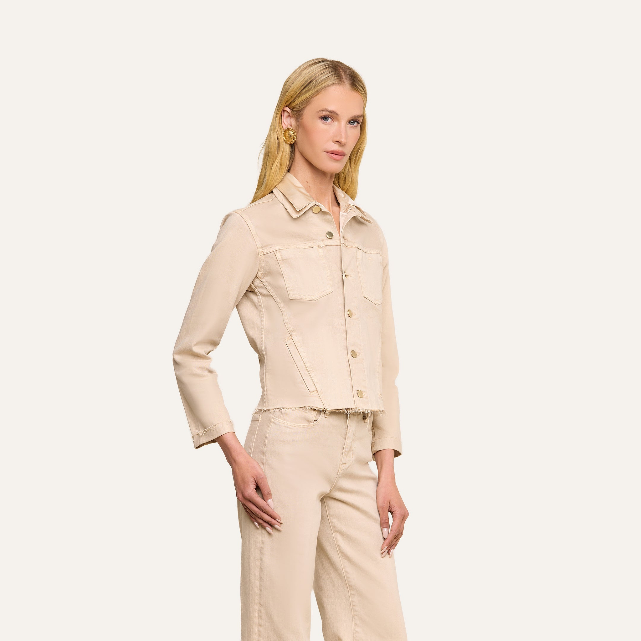Une femme aux longs cheveux blonds porte la veste « Janelle Jean » de couleur beige avoine et un pantalon taille haute assorti, devant un fond beige uni ; elle a une main le long du corps et lautre sur la hanche.