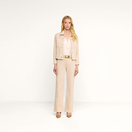 Une femme aux longs cheveux blonds se tient debout devant un fond beige. Elle porte la veste « Janelle Jean » de couleur beige avoine, associée à un chemisier blanc, un pantalon beige à jambes larges, une ceinture marron et des talons. Son expression est neutre.