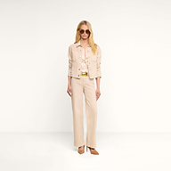 Une femme aux longs cheveux blonds pose devant un fond beige, vêtue de lunettes de soleil, dune veste « Janelle Jean » de couleur beige avoine à la coupe féminine, dun pantalon large de couleur crème, dun chemisier soyeux de couleur crème, dune ceinture dorée et de talons hauts couleur beige clair.