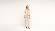 A woman wearing sunglasses and an all-beige outfit, including a jacket and wide-leg pants, stands against a plain white background.