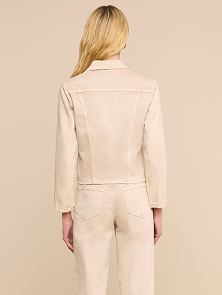 Eine Frau mit langen blonden Haaren steht mit dem Rücken zur Kamera und trägt die Janelle-Jeansjacke in Oatmeal-Beige sowie eine dazu passende Stretch-Jeanshose vor einem schlichten beigen Hintergrund.
