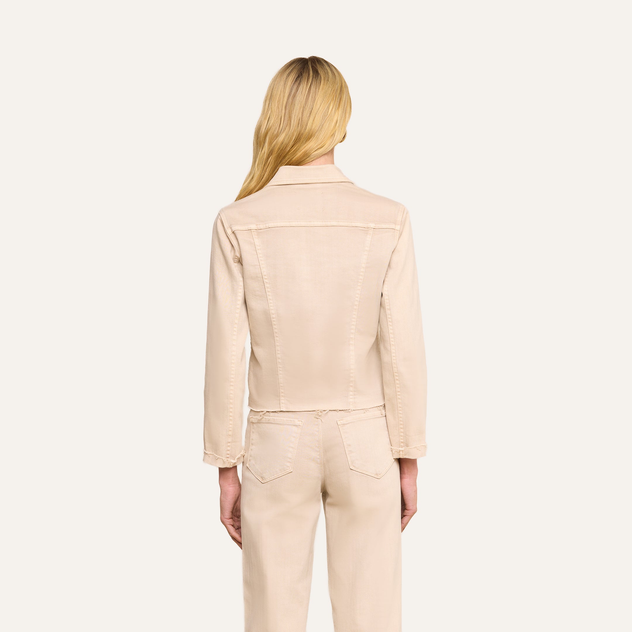 Une femme aux longs cheveux blonds, de dos, porte la veste « Janelle Jean » de couleur beige avoine, assortie à un pantalon en denim stretch de la même teinte, sur un fond beige uni.