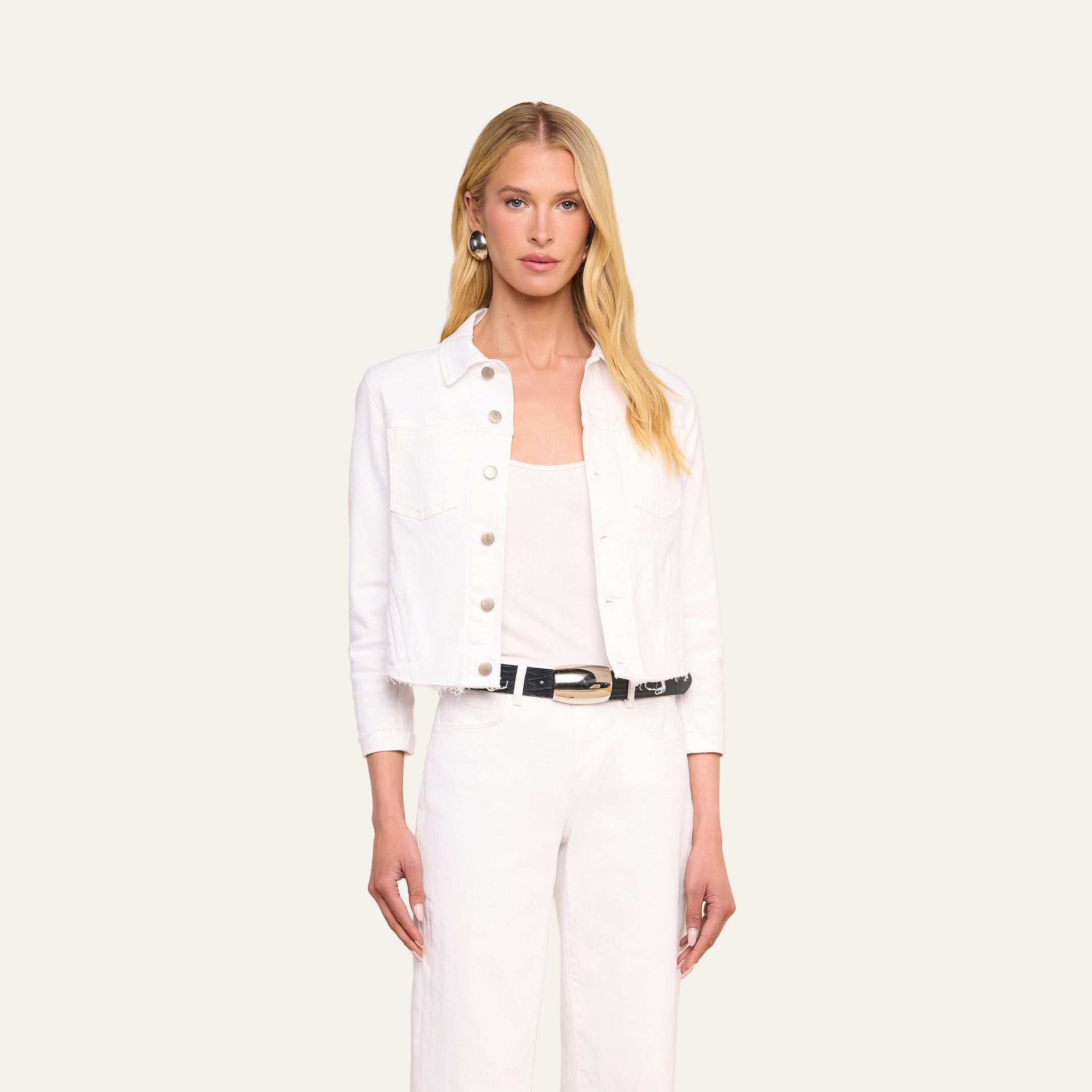 Indossando la giacca “Janelle Jean”, una donna dai lunghi capelli biondi abbina questo capo in denim elasticizzato bianco dal taglio femminile a un top bianco e a pantaloni bianchi, completando il look con una cintura nera, su uno sfondo beige.